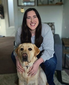 Kali O'Meara smiling with long straight dark brown hair and wearing a gray top and jeans. She is sitting on a chair with a yellow lab dog sitting in front of her.