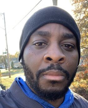 Dr. Vignon Oussa with black mustache and beard wearing a black beanie, earbuds and a blue shirt under a gray jacket
