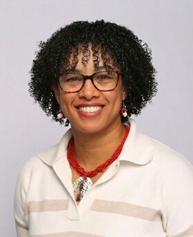 Cleonie Mainvielle smiling with medium length curly black hair and brown rim glasses and wearing a white and beige v-neck sweater