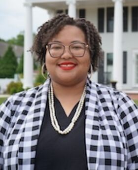 Dr. Sheena Rancher stands outside of the MRISJ office at 161 Summer street. She has short, curly hair and is wearing glasses with large earrings and a draped pearl necklace. Her blazer is black and white gingham