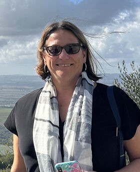 Dr. Gia Renaud smiling with medium length straight brown hair and wearing sunglasses, a short sleeve black top and a white and black scarf