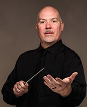 Dr. Donald Running wearing a black button down shirt with black tie, holding a conductor's wand in one hand while his other hand is held out towards the camera