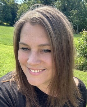 Maryjo Sabalewski smiling with medium length straight brown hair and wearing a black top. Grass and trees are in the background.