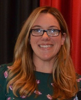 Dr. Melinda Tarsi-Goldfien smiling wearing black rim glasses, long dirty blonde wavy hair and a dark green top with pink floral pattern