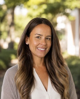 Dr. Hannah Taverna smiling with long brown wavy hair and wearing a gray open sweater over a white v-neck blouse