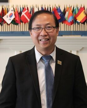Dr. Wing-Kai To smiling with short graying black hair, wearing black rimmed glasses and a black suit jacket over a white button down shirt with a blue tie