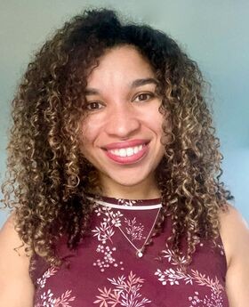 Dr. Maggie Veiga smiling with medium long curly dark brown hair with highlights and wearing a sleeveless maroon top with white and pink leaves on it