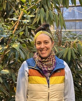 Emma Welch standing in a greenhouse smiling with brown hair in a messy bun on top of her head wearing a gold headband, gray long sleeve sweatshirt over an LLBean puffy vest with blue collar and orange, gold and beige stripes. She also wears 2 fashion scarves in colors of purple, gold and orange