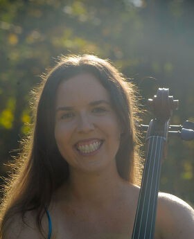 Dr. Sara Wilkins smiling with sun shining on her long dark brown hair. She is wearing a blue spaghetti strap top and the neck of her cello is in front of her shoulder