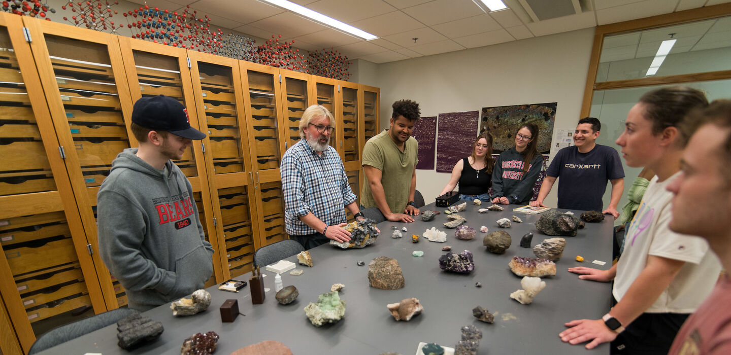 Dr. Michael Krol teaches class while eight students stand around the table with him. The table has several different kinds of rocks laid out on it.