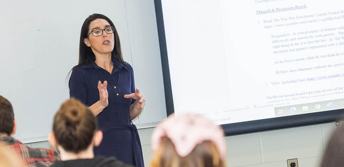 Professor Ashley Rodrigues teaching at the front of the class next to a large screen with course material projected on it while students look on in the foreground
