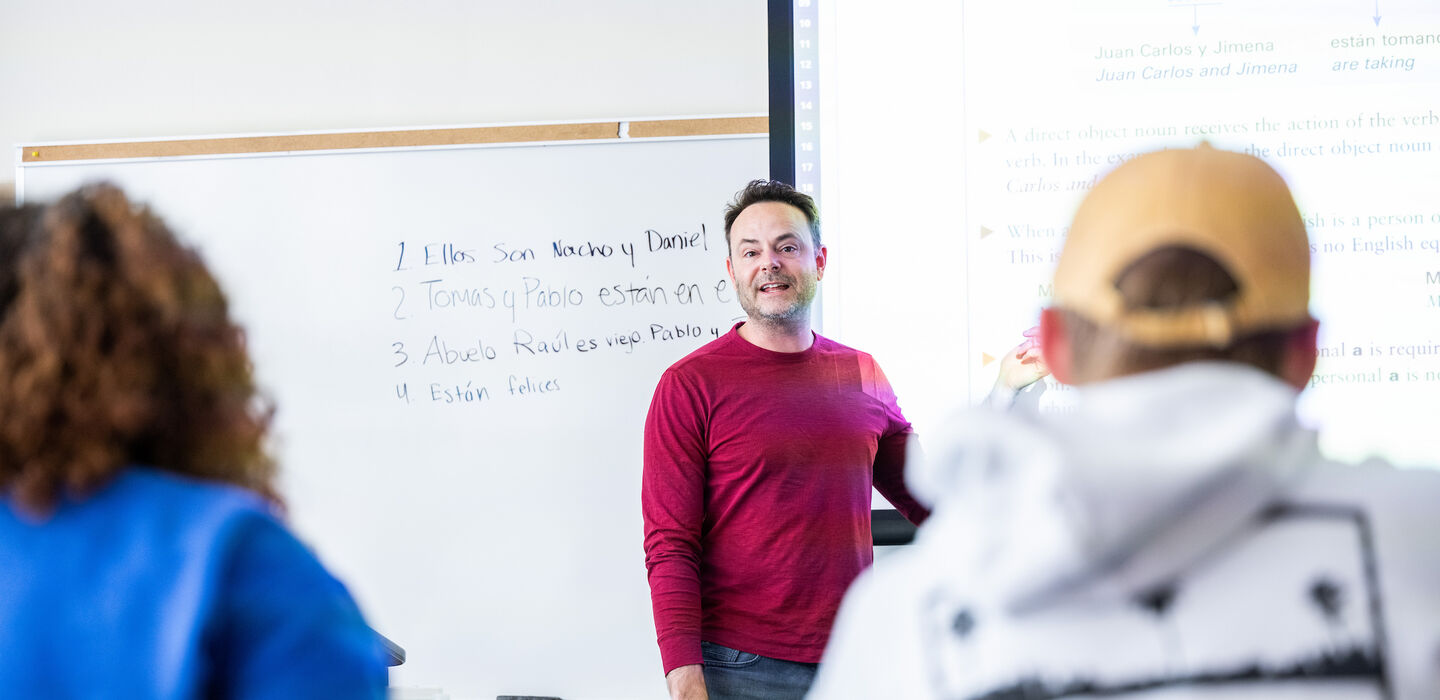 looking over the shoulders of two students at Dr. Ryan LaBrozzi teaching a Spanish class in front of a white board and projector screen 