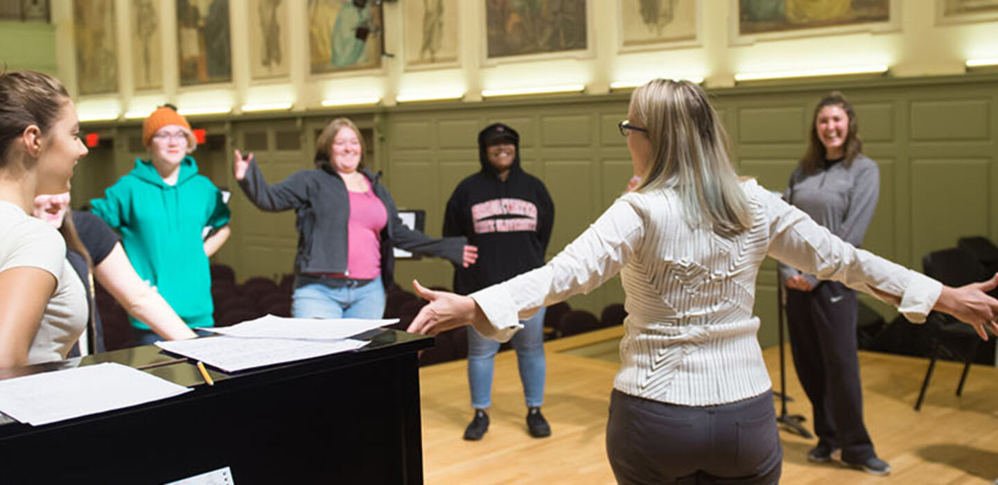 BSU music professor stands next to a piano on stage with her back to the camera and her arms open wide as part of a circle of music students