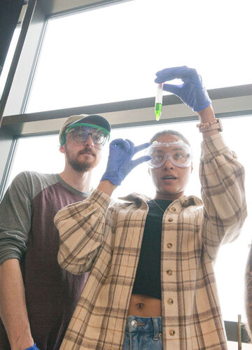 a student holds a test tube with green liquid up to the light while another student looks on